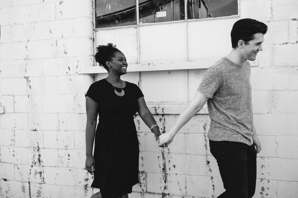 Black-and-white photo of a couple holding hands and smiling while walking beside an industrial brick wall. 